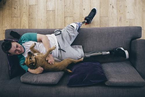 Amputee teen boy on couch with dog laughing.