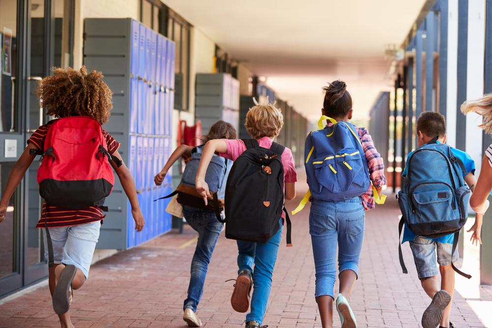 Diverse group of kids running down a school hallway with backpacks on.