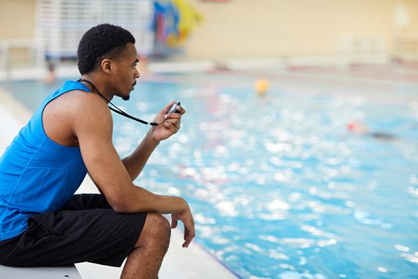 Black male swim coach timing swimmers in indoor pool.