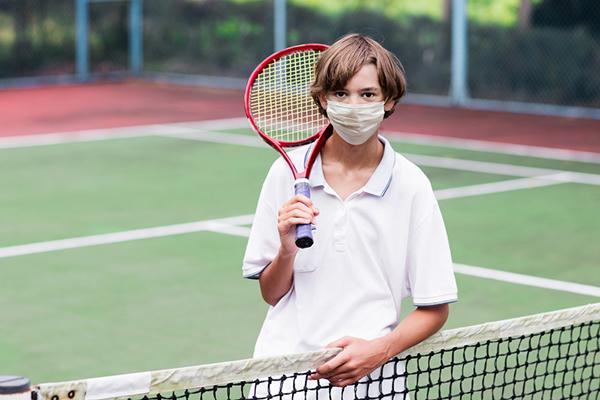 Young white boy holding a tennis racket with a mask over his nose and mouth.