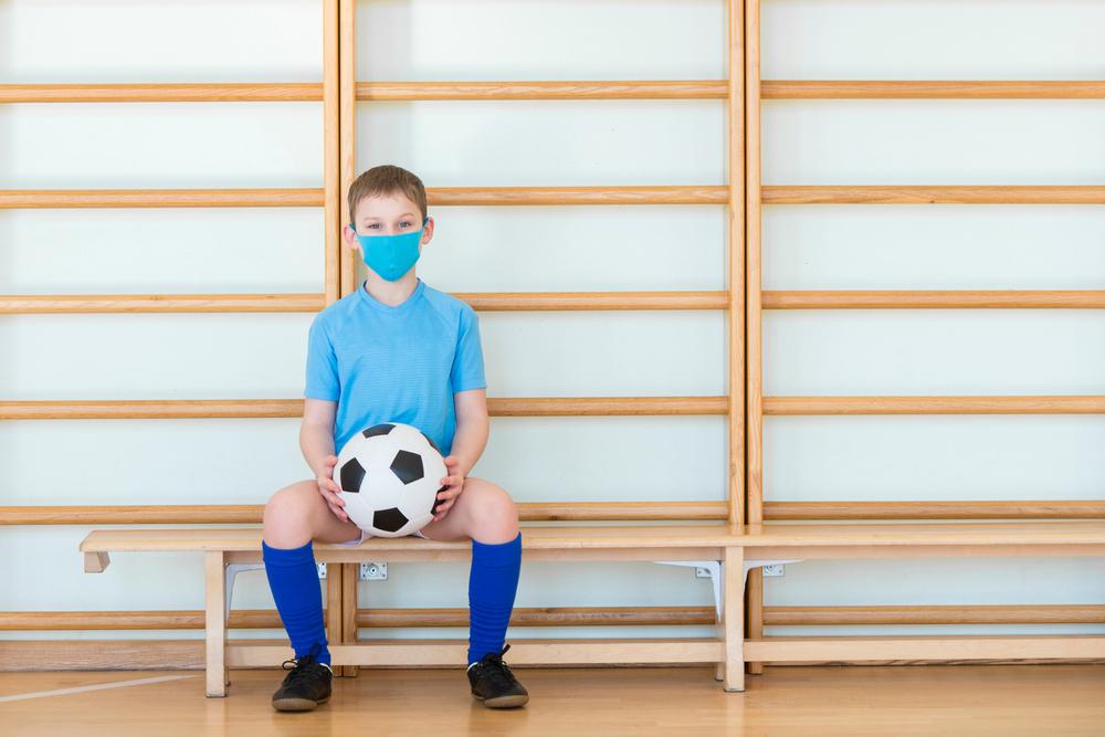 Young boy sitting on indoor bench wearing a mask and holding a soccer ball.