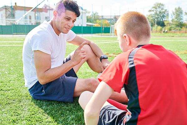 Male coach talking to a young male soccer player while sitting on the field.