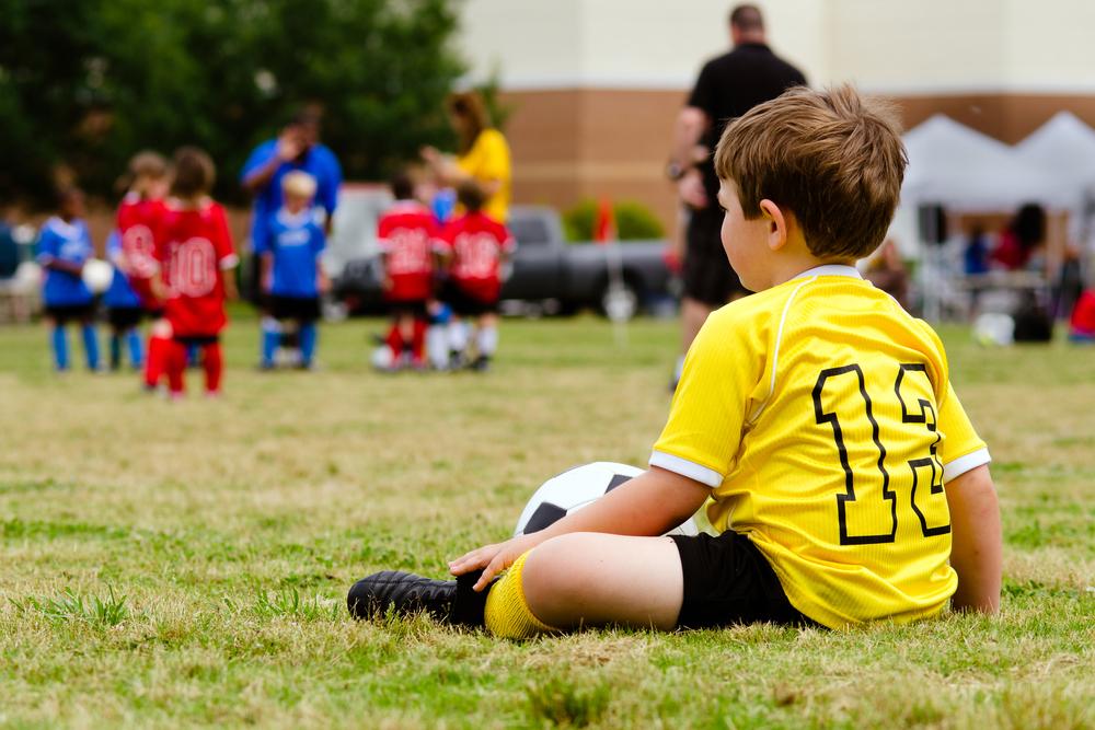 Young child sitting on sidelines watching teammates play soccer.