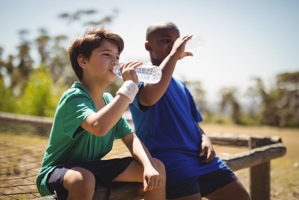 Two young boys drinking water out of water bottles on a bench outside.