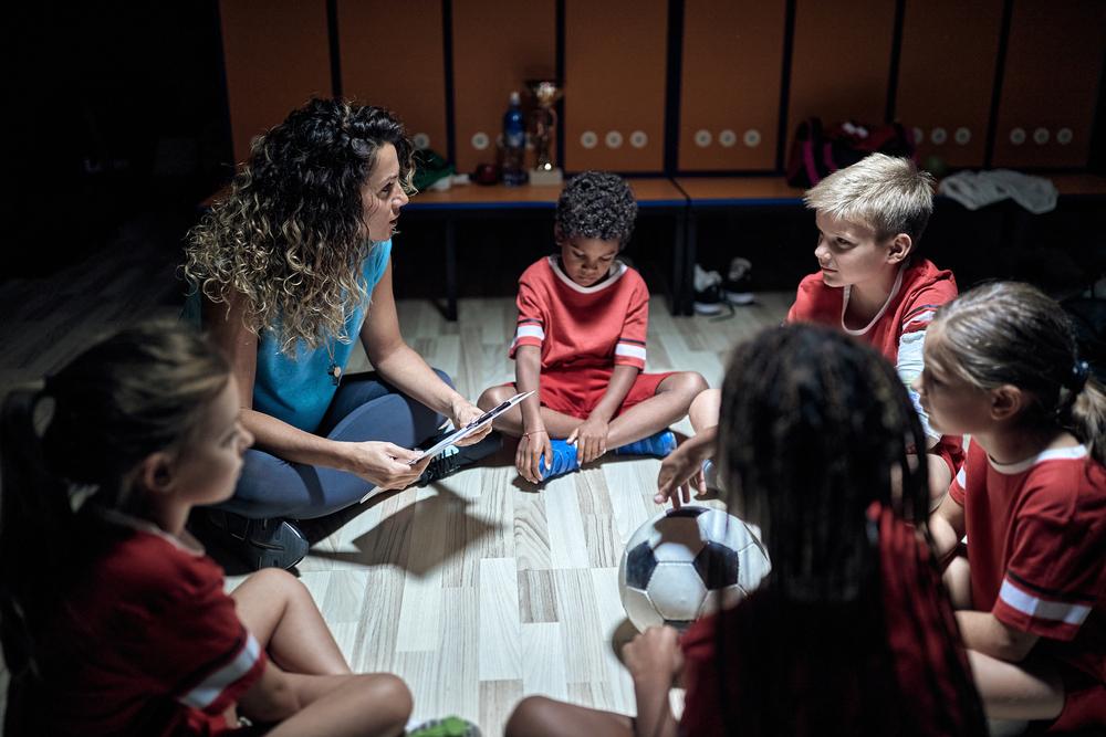 Coach sitting with young soccer team in locker room.