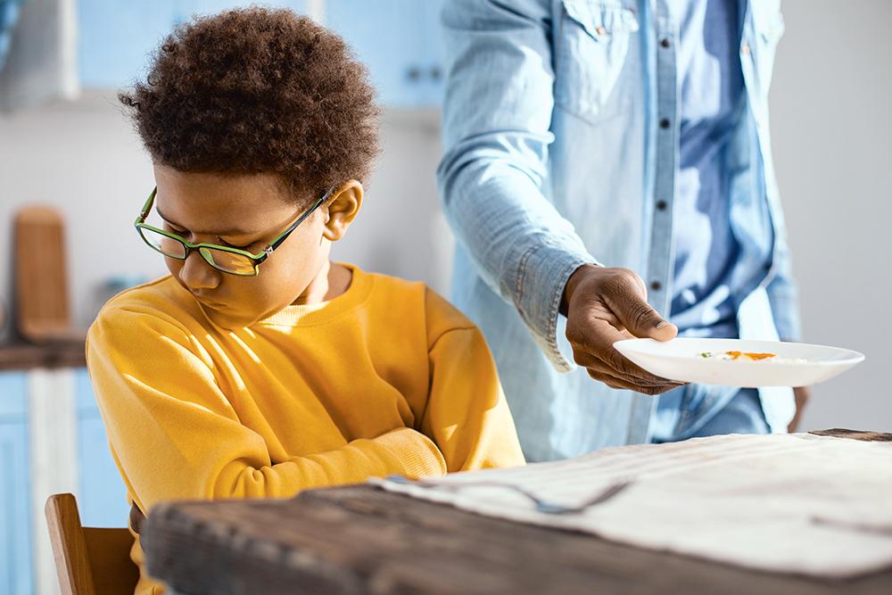 Young black child turning away from a plate of food handed to him by a parent.