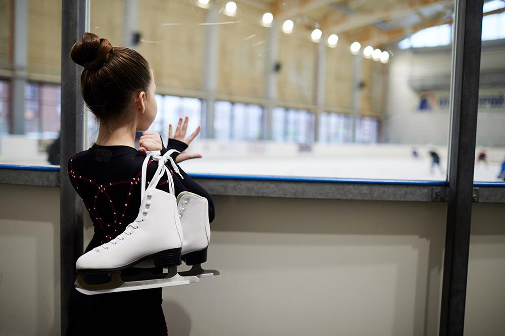 White girl looking at skating rink while holding figure skating skates.