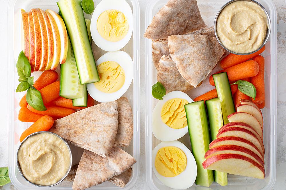 Two tupperware containers filled with veggies, hard boiled eggs, pita, hummus, and sliced apples.