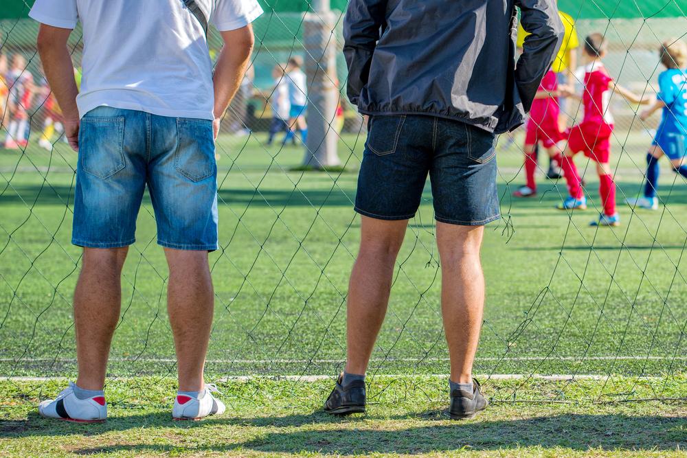 Rear view of two dads on sidelines at youth sports game.