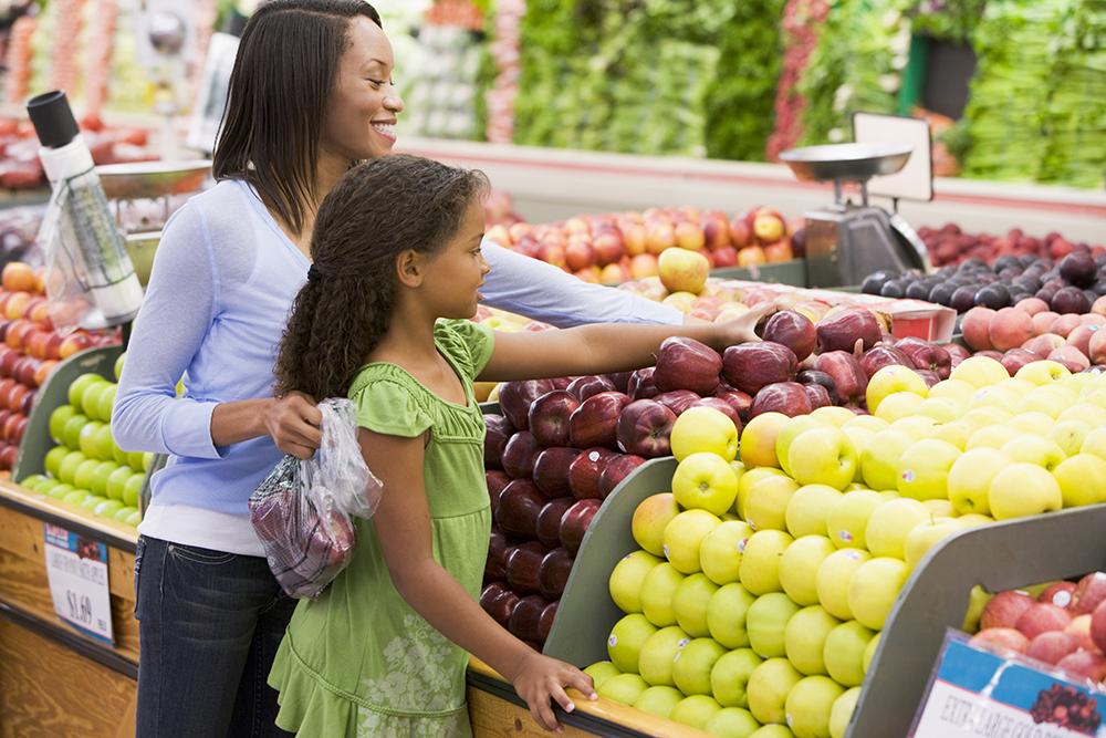 Black mom helping daughter grab apple at a grocery store.