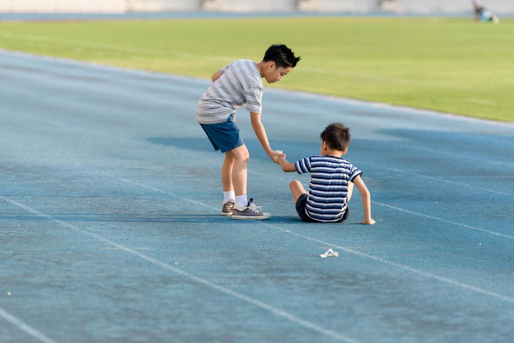 Young boy helping another younger boy stand up on a track.