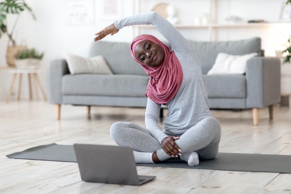 Young black woman wearing a hijab stretching in front of a laptop in her home.